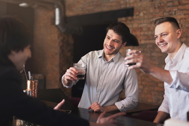 Handsome smiling friends drinking whiskey and communicating with cheerful bartender in pub, copy space