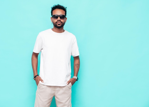 Handsome smiling hipster model Sexy unshaven man dressed in white summer tshirt and jeans clothes Fashion male posing near blue wall in studio Isolated In sunglasses