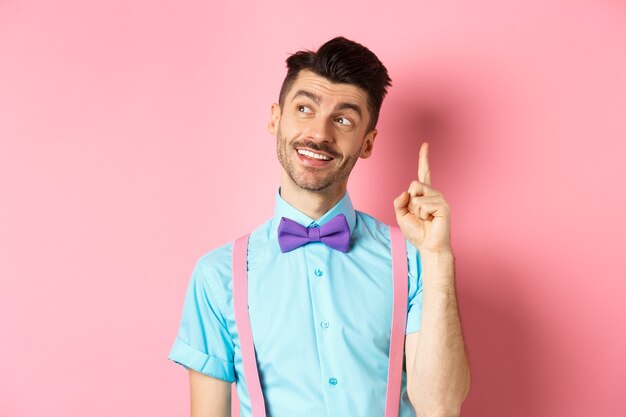 Handsome smiling man raising finger in eureka gesture, looking away and pitching an idea, have suggestion or plan, standing in bow-tie over pink.