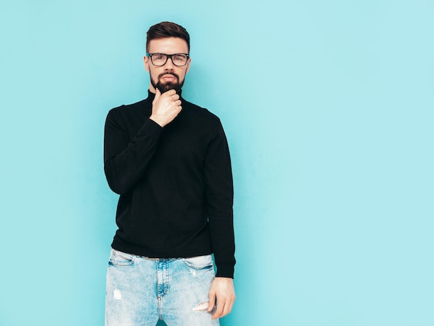 Handsome smiling model Sexy stylish man dressed in black turtleneck sweater and jeans Fashion hipster male posing near blue wall in studio Isolated