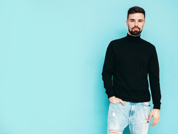 Handsome smiling model Sexy stylish man dressed in black turtleneck sweater and jeans Fashion hipster male posing near blue wall in studio Isolated