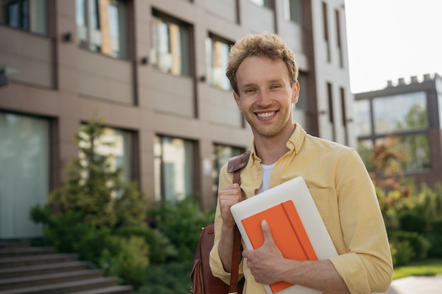 Handsome smiling student wearing casual clothing holding laptop and book looking at camera