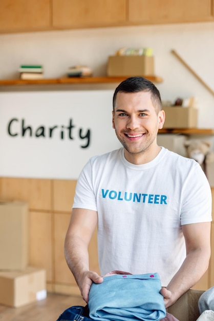 Handsome smiling volunteer holding clothes and looking at camera in charity center