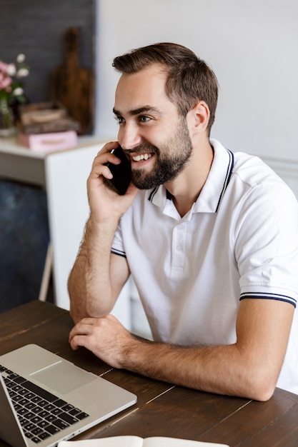 Handsome smiling young bearded man sitting at the table at home, using laptop computer, talking on mobile phone