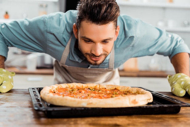 Handsome smiling young man smelling fresh homemade pizza on baking tray