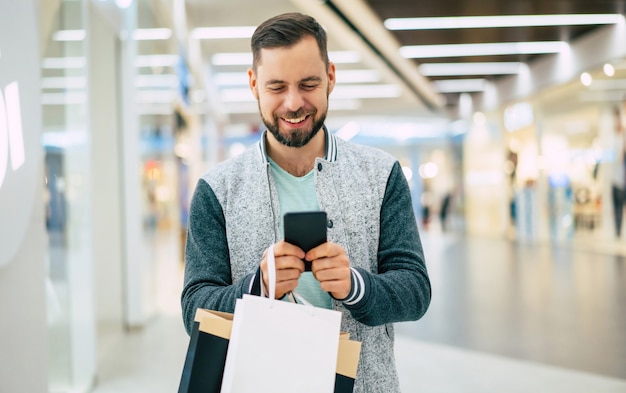 Handsome smiling young stylish bearded man with shopping bags is using smart phone while walking in the mall 