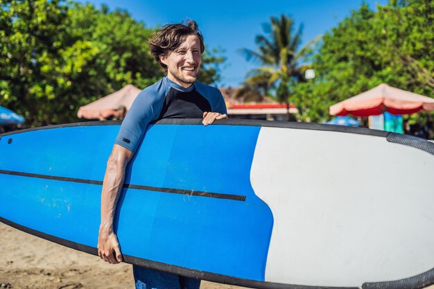 Handsome sporty young surfer posing with his surfboard under his arm in his wetsuit on a sandy tropical beach