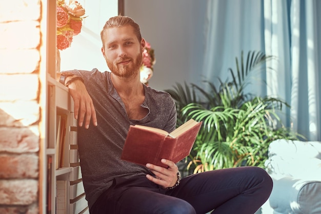 A handsome stylish redhead student in casual clothes, sitting on a chair at a home and holds a book, looking at a camera.