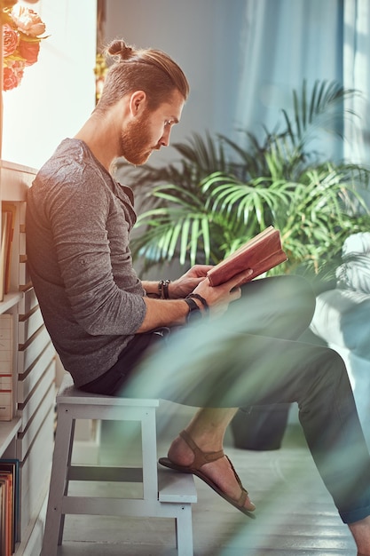 A handsome stylish redhead student in casual clothes, sitting on a chair at a home while reading a book.