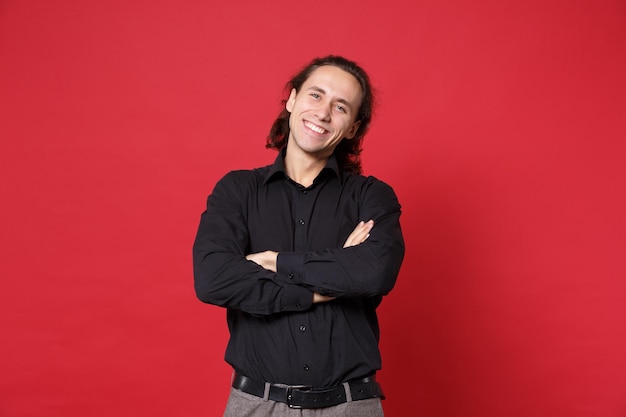 Handsome stylish young curly long haired man in black shirt posing isolated on red wall background studio portrait. People sincere emotions lifestyle concept. Mock up copy space. Holding hands crossed