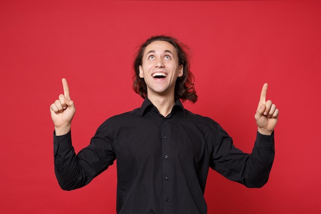 Handsome stylish young curly long haired man in black shirt posing isolated on red wall background studio portrait. People sincere emotions lifestyle concept Mock up pointing finger hand on copy space