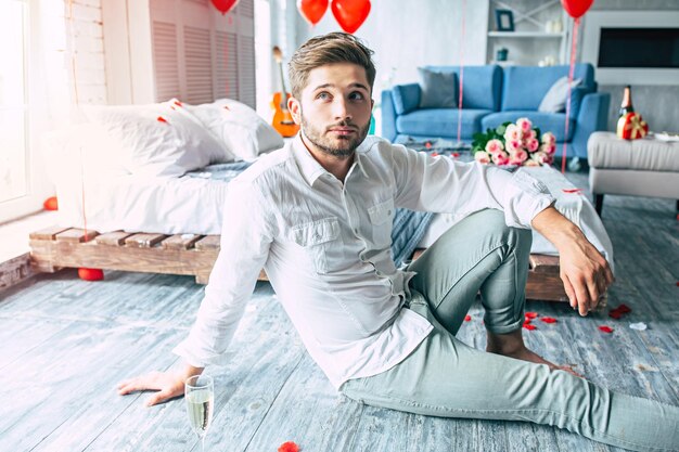 Handsome stylish young man is sitting on floor on romantic bedroom background while he is waiting on his beloved