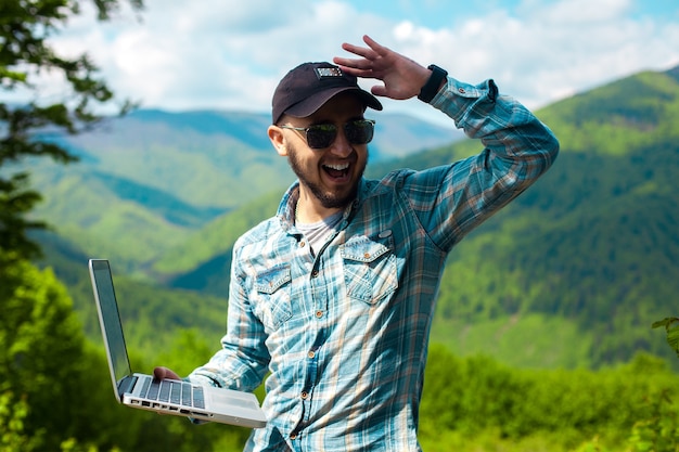 Handsome stylish young man in sunglasses and with a laptop in hands looking aside and smiling broadly in the mountains