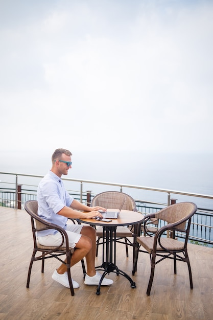 Handsome successful young male businessman sitting at a table by the pool with a laptop overlooking the Mediterranean Sea. Remote work on vacation. Vacation concept