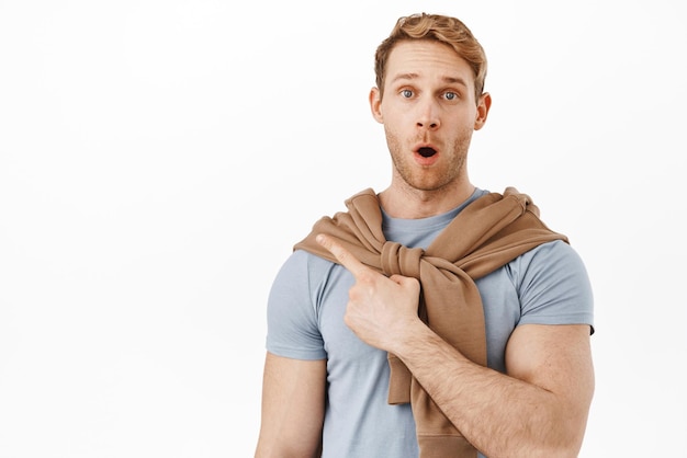 Handsome surprised redhead man gasping say wow and pointing at upper left corner item showing product logo or banner impressed by promotional text standing over white background