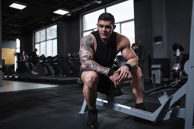 Handsome tattooed muscular bodybuilder resting at the gym after exercising