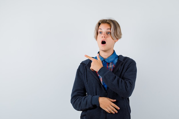 Handsome teen boy in shirt, hoodie pointing left, looking away and looking shocked , front view.