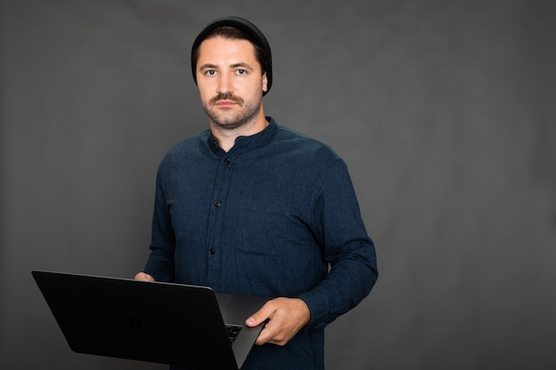 Handsome unshaven guy in knitted cap posing with laptop on gray studio