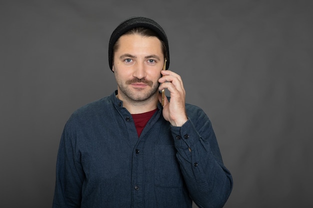 Handsome unshaven guy in knitted cap talking on phone on gray studio