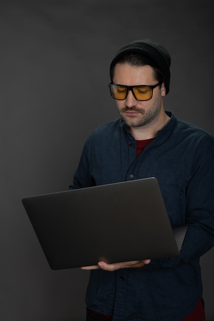 Handsome unshaven hipster in knitted cap working with laptop on gray backdrop