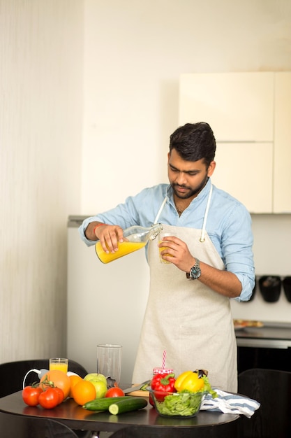 Handsome unshaven indian man wearing blue shirt and apron pouring fresh organic orange juice into a glass looking at camera, prepearing breakfast in kitchen at home. Healthy vegan lifestyle.