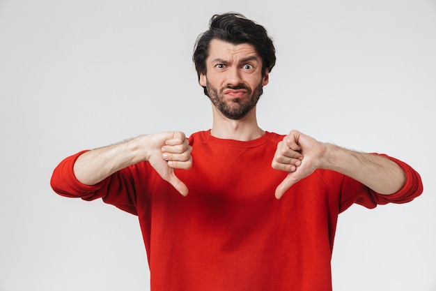 Handsome upset young bearded brunette man wearing sweater standing over white, thumbs down