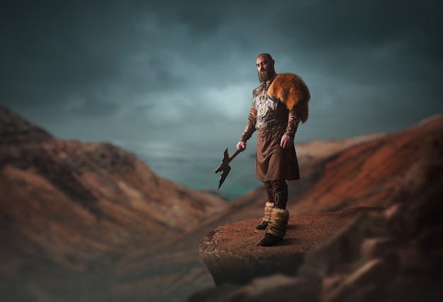 Handsome viking with axe dressed in traditional nordic clothes standing on the top of rocky mountain. Scandinavian ancient warrior