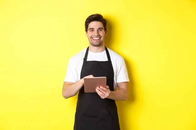 Handsome waiter taking orders, holding digital tablet and smiling, wearing black apron uniform, standing over yellow wall