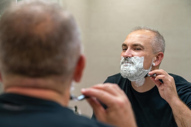 Handsome whitehaired beared man shaving off his beard looking in a mirror Concept of changing style