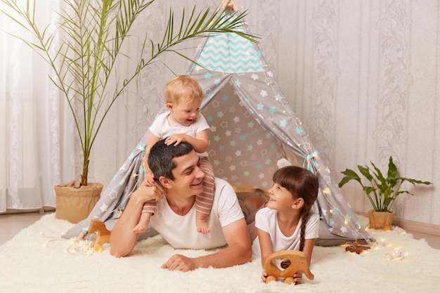 Handsome young adult father with little daughters having fun and playing in peetee tent in children room at home infant baby sitting on dad's back family with happy expressions