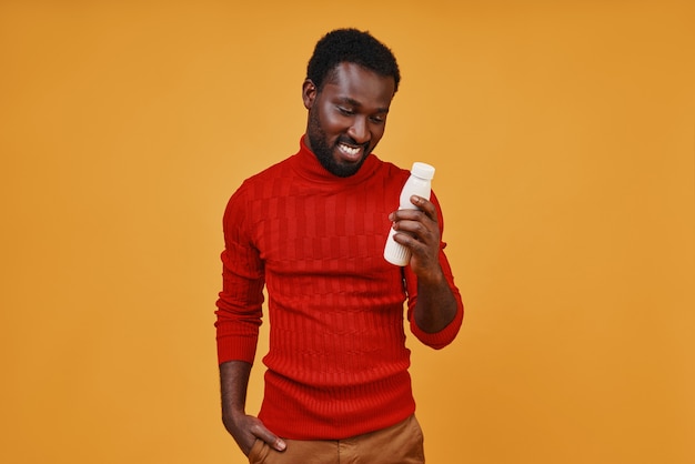 Handsome young African man looking at the bottle and smiling while standing against yellow background