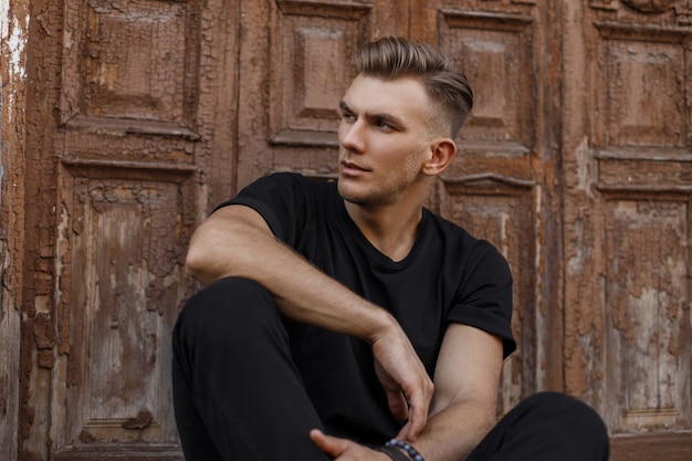 Handsome young american model man with hairstyle in black trendy t-shirt sitting near a vintage wooden door