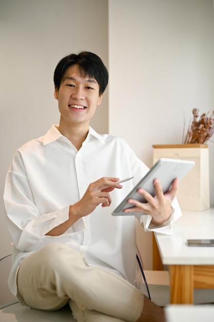 A handsome young Asian man sits in a minimal bright coffee shop with his digital tablet