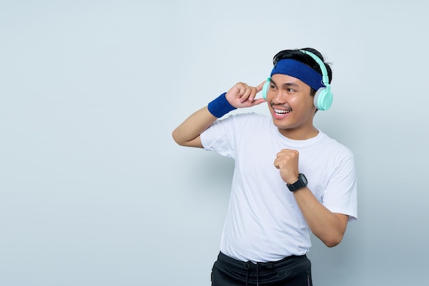 Handsome young asian man sporty fitness trainer instructor in blue headband and white tshirt Makes fun gesture while Listening to music with headphones isolated over white background