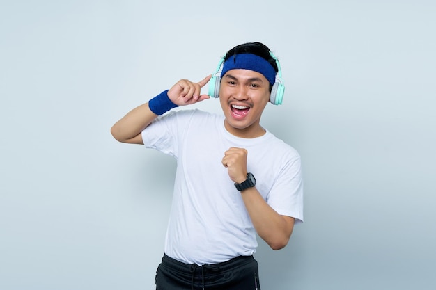 Handsome young asian man sporty fitness trainer instructor in blue headband and white tshirt Makes fun gesture while Listening to music with headphones isolated over white background