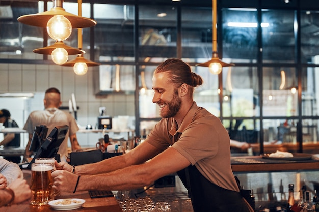 Handsome young bartender in apron serving beer and smiling while working in the pub