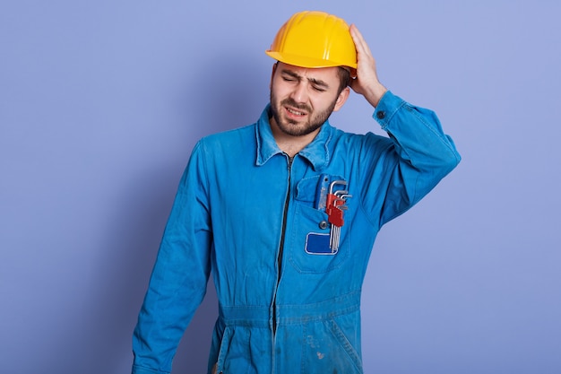 handsome young bearded Caucasian mechanic wearing blue work clothes and yellow hard hat, stands touching his head