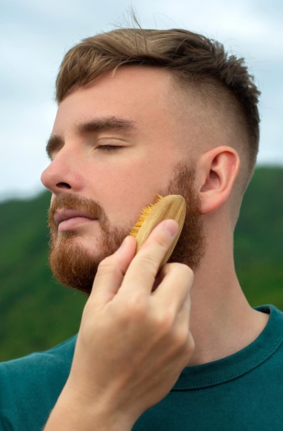 Handsome young bearded guy brutal man with a beard hair is combing the beard on face with a comb brush Male personal self care products accessory Natural summer background vertical photo