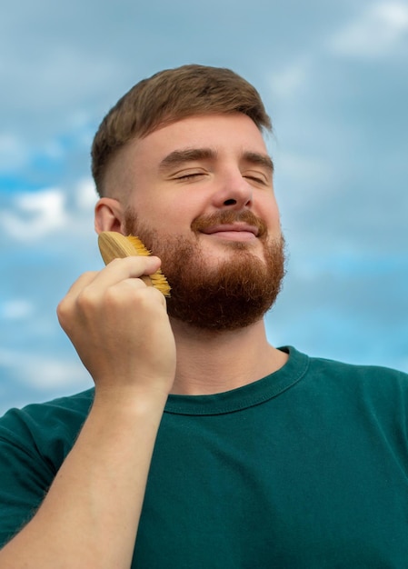 Handsome young bearded guy brutal man with a beard hair is combing the beard on face with a comb brush Male personal self care products accessory Natural summer background vertical photo