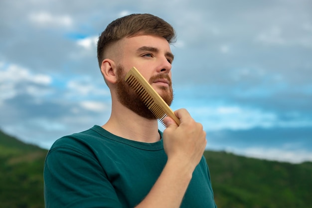 Handsome young bearded guy European brutal man with a beard hair is combing the beard on face with a comb brush Male personal self care products accessory on nature Natural summer background