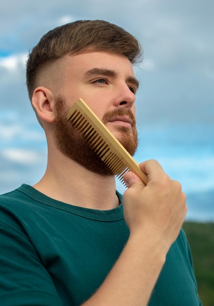 Handsome young bearded guy man with a beard hair is combing the beard on face with a comb brush Male personal self care products accessory on nature Natural summer background Vertical photo