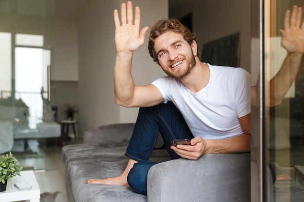  a handsome young bearded man on sofa at home waving to window using mobile phone.