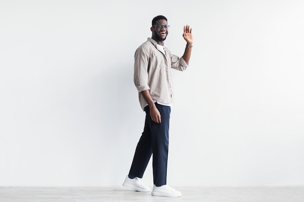 Handsome young black man walking and waving hand at camera greeting someone against white wall copy