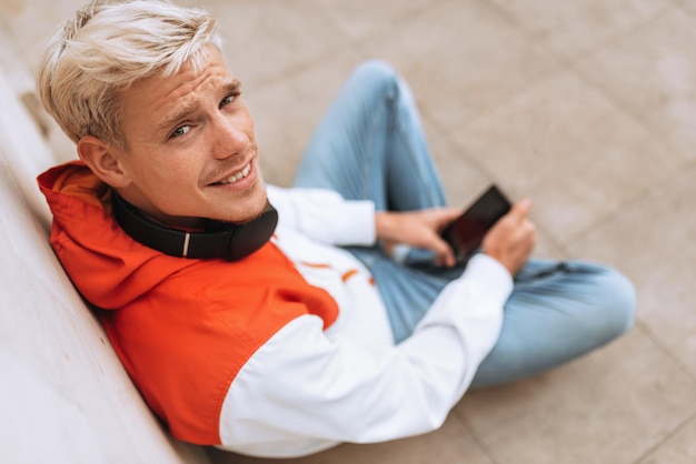 Handsome young blond man enjoying weather in city park waiting for his girlfriend texting message while listening to his favorite music from wireless headphone using smart phone outdoor in the park