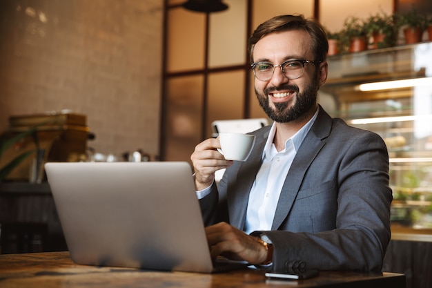 Handsome young businessman dressed in suit working on a laptop computer, sitting at the cafe indoors, drinking coffee