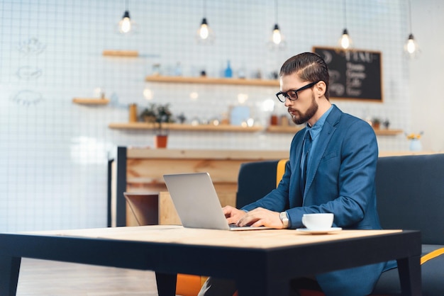 Handsome young businessman in eyeglasses using laptop while drinking coffee in cafe