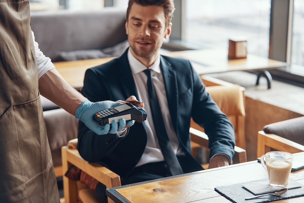 Handsome young businessman making a contactless payment while sitting in the restaurant