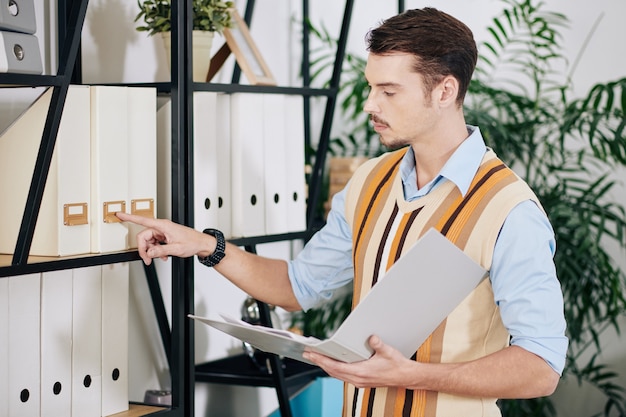 Handsome young businessman with folder in hands searching for document in office archive