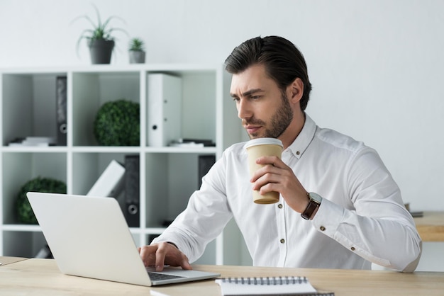 Handsome young businessman with paper cup of coffee working with laptop