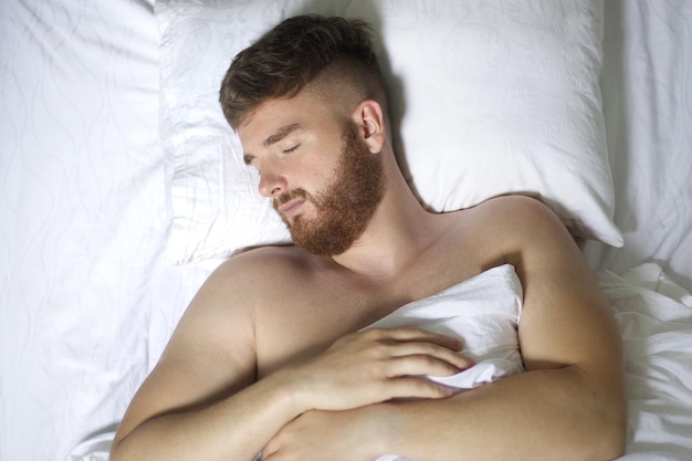 Handsome young calm sleepy bearded man with beard is sleeping well on side in bed in bedroom on pillow with his eyes closed in the morning resting Good healthy dream rest White linens Top view